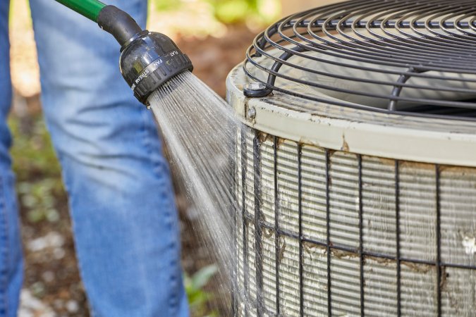 Woman uses garden hose to rinse debris from air conditioner compressor.