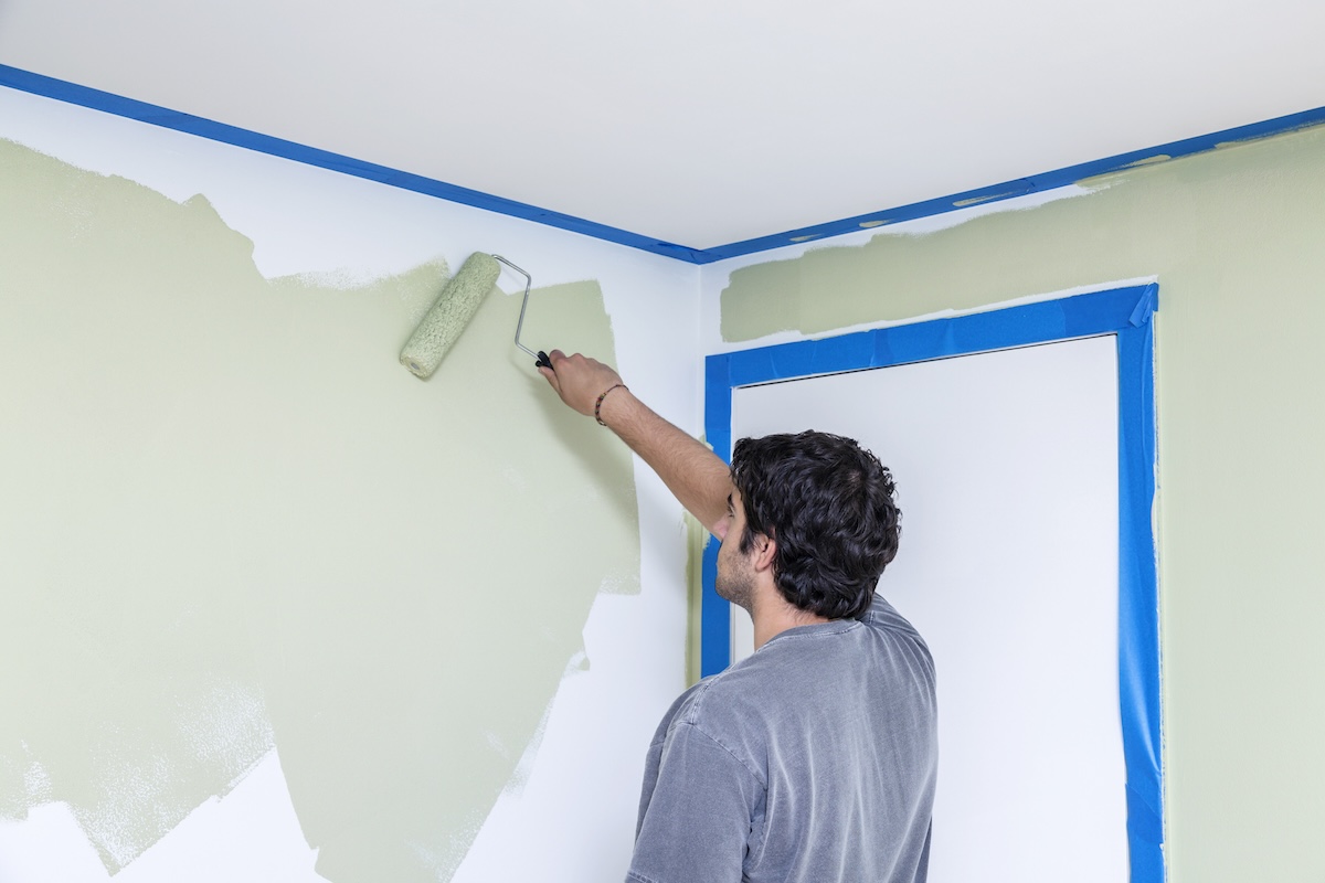 A man in a grey shirt paints a wall light green with painter's tape.