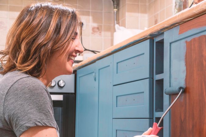 Woman sitting on her kitchen floor, painting her brown lower kitchen cabinets blue.