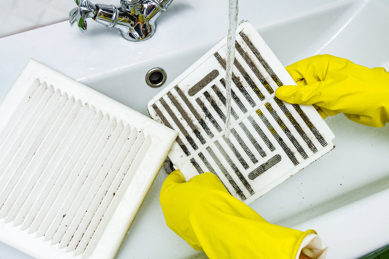Person wearing yellow rubber gloves rinses a bathroom exhaust fan cover and filter in sink.